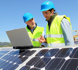 Solar workers wearing safety vests and hard hats looking at a laptop by solar panels.